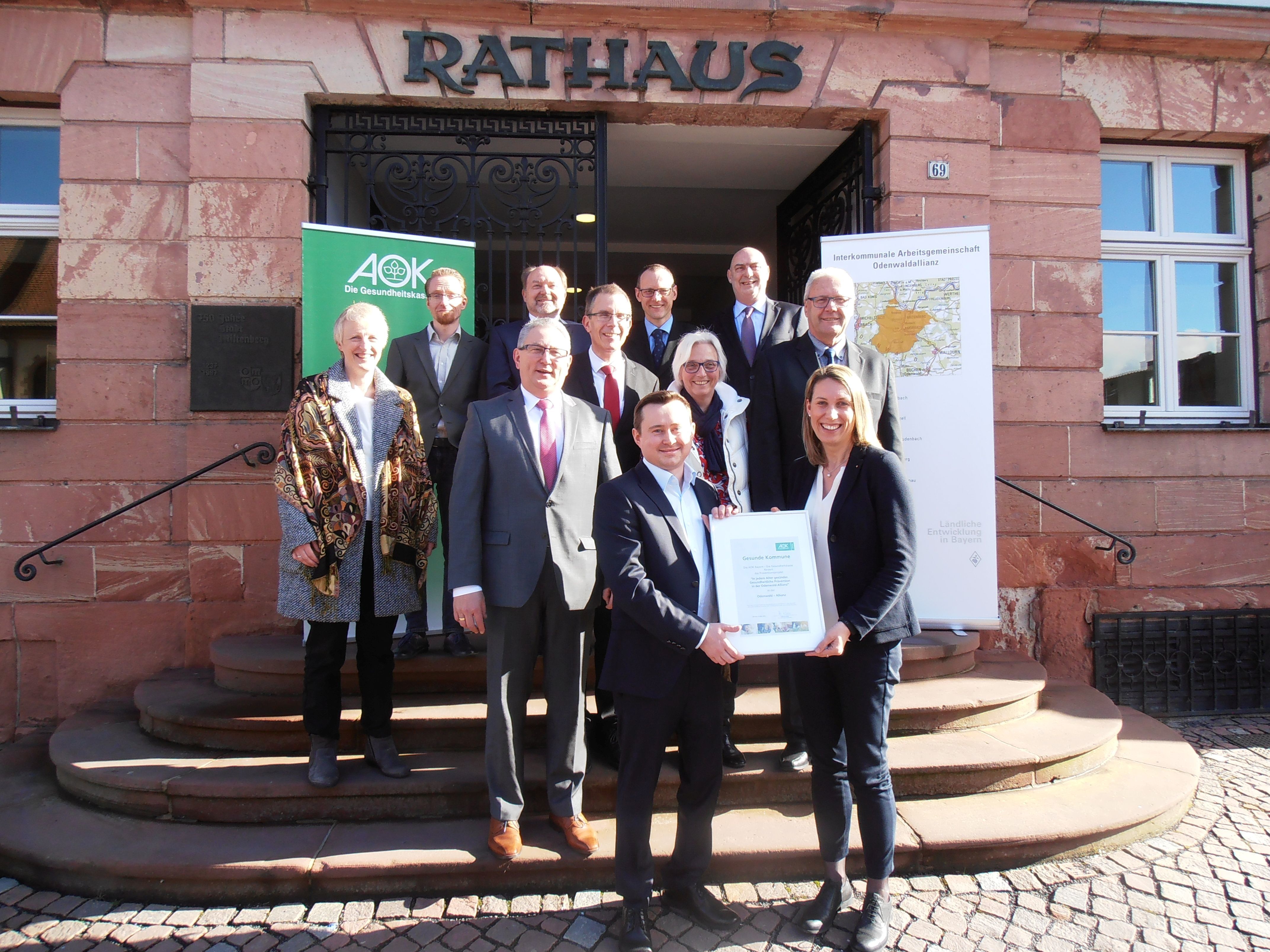 Frau Daniela Bergmann, Direktorin der AOK Bayern in Aschaffenburg überreichte die Urkunde für die Odenwald – Allianz an Allianzmanager Viktor Gaub (vorne).
Zweite Reihe von links: Bürgermeister Peter Schmitt (Amorbach), zweite Bürgermeisterin Susanne Wörner (Kirchzell), Bürgermeister Bernhard Kern (Weilbach)
Dritte Reihe von links: Christine Becker (SalutoConsult), Prof. Dr. Jürgen Zerth (WLH)
Letzte Reihe von links: Sebastian Müller (WLH), Bürgermeister Helmut Demel (Miltenberg) , Bürgermeister Udo Käsmann (Rüdenau), Bürgermeister Bernd Klein (Laudenbach)