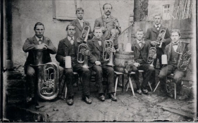 Die Fotoaufnahme aus dem Jahr 1900 zeigt die sieben Gründungsmitglieder der ersten Mechenharder Musikkapelle zusammen mit ihrem Ausbilder (von links nach rechts): Ludwig Bernard, Leo Bernard, Joseph Rüttiger, Alois Berninger, Musiklehrer Otto Miltenberger (mit Geige) aus Rück, Anton Heider, Philipp Vill und Ignaz Berninger. Das Gruppenbild entstand, wie der Chronist Franz Heider berichtet, nachdem ein edler Spender den Musikern nach ihrem ersten Auftritt zur Wallfahrt nach Röllbach ein Fass Bier gestiftet hatte.