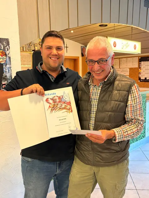 Der 1. Vorsitzende Alexander Fäth, links im Bild, überreichte die goldene Ehrennadel des BTTV für 40 Jahre aktiven Tischtennissport an Konrad Fäth, rechts im Bild. Beide Männer stehen, lachen und halten die Ehrenabzeichen in die Kamera.