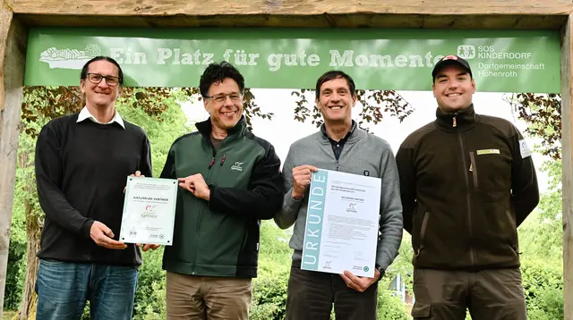 Vier Männer stehen nebeneinander im Grünen und halten eine Urkunde und Plakette hoch.