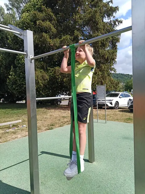 Ein Kind probiert an der Calisthenics-Anlage erste Klimmzüge aus und nutzt dabei ein Widerstandsband als Einstiegshilfe.
