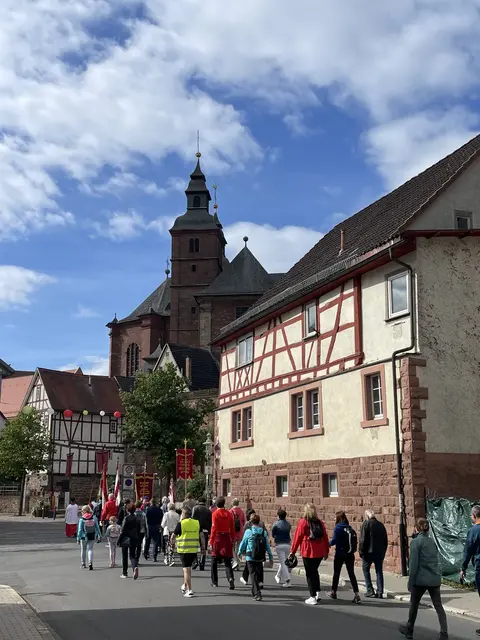 
Gnadenweg, Glaubensschritte, Gemeinschaft – Wallfahrt im Zeichen der Liebe.


Die Walldürn‑Wallfahrt 2026 schrieb sich am 14. Juni in die frühen Stunden des Sonntags, als die Pilgerinnen und Pilger der Pfarreiengemeinschaften Miltenberg‑Bürgstadt und Eichenbühl‑Erftal im ersten Licht aufbrachen – geführt vom Leitwort „Die Liebe hört niemals auf“, das wie ein stiller Kompass über dem ganzen Tag lag. Viele gingen aus Dankbarkeit, andere aus Bitte, wieder andere aus der Sehnsucht nach Orientierung; manche suchten Trost, manche wollten ein Versprechen erneuern, manche einfach spüren, dass Glauben im Gehen Gestalt annimmt.

Die Musikerinnen und Musiker aus Miltenberg, Schippach, Wenschdorf, Monbrunn, Eichenbühl und Neunkirchen gaben diesem Weg einen warmen Atem. Ihre Bläserklänge legten sich wie ein tragender Teppich unter die anstrengenden Kilometer, verwandelten Steigungen in Rhythmus und Pausen in kleine Inseln der Sammlung. In Gerolzahn und Glashofen mischten sich Stimmen, Gebete und Erinnerungen – Momente, in denen Gemeinschaft nicht erklärt, sondern erlebt wurde.

Der Einzug in Walldürn wurde durch die Musik zu einem Augenblick, der Ankunft und Aufbruch zugleich war. In der Basilika füllte der Pontifikalgottesdienst mit Erzbischof Dr. Ludwig Schick den Raum mit einer zeitgemäßen Predigt, die das Jubiläum „400 Jahre Blutaltar“ mit den Hoffnungen der Gegenwart verband. Viele Pilger spürten, dass dieser Ort nicht nur Geschichte bewahrt, sondern Herzen sammelt und stärkt.

Nach der Verabschiedung an der Gnadenstätte begann die Rückkehr – stiller, aber erfüllt von dem Gefühl, etwas Wertvolles mitgenommen zu haben. Das ideale Wetter machte den Weg leicht, ohne die Ernsthaftigkeit des Pilgerns zu mindern. So bleibt diese Wallfahrt als ein Tag in Erinnerung, an dem Menschen suchten, fanden und getragen wurden.

Text und Foto: Roland Schönmüller






